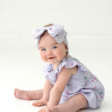 A smiling blue-eyed baby sits on the floor amid pink tulips, wearing the Mabel and Honey Tiny Tulip Baby Romper with ruffles and a matching cherry-print bow headband, set against a white background.