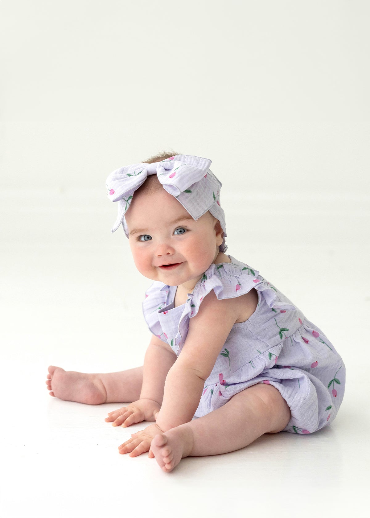 A smiling blue-eyed baby sits on the floor amid pink tulips, wearing the Mabel and Honey Tiny Tulip Baby Romper with ruffles and a matching cherry-print bow headband, set against a white background.