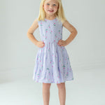 A young blonde girl smiles with her hands on her hips, wearing the Mabel and Honey Tiny Tulip Drop Waist Dress and silver sandals, standing against a plain white background.