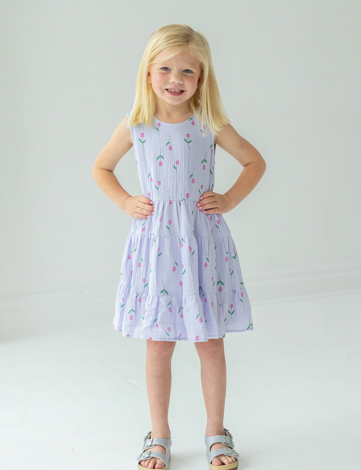 A young blonde girl smiles with her hands on her hips, wearing the Mabel and Honey Tiny Tulip Drop Waist Dress and silver sandals, standing against a plain white background.