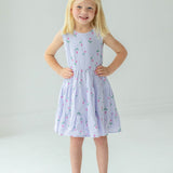 A young blonde girl smiles with her hands on her hips, wearing the Mabel and Honey Tiny Tulip Drop Waist Dress and silver sandals, standing against a plain white background.