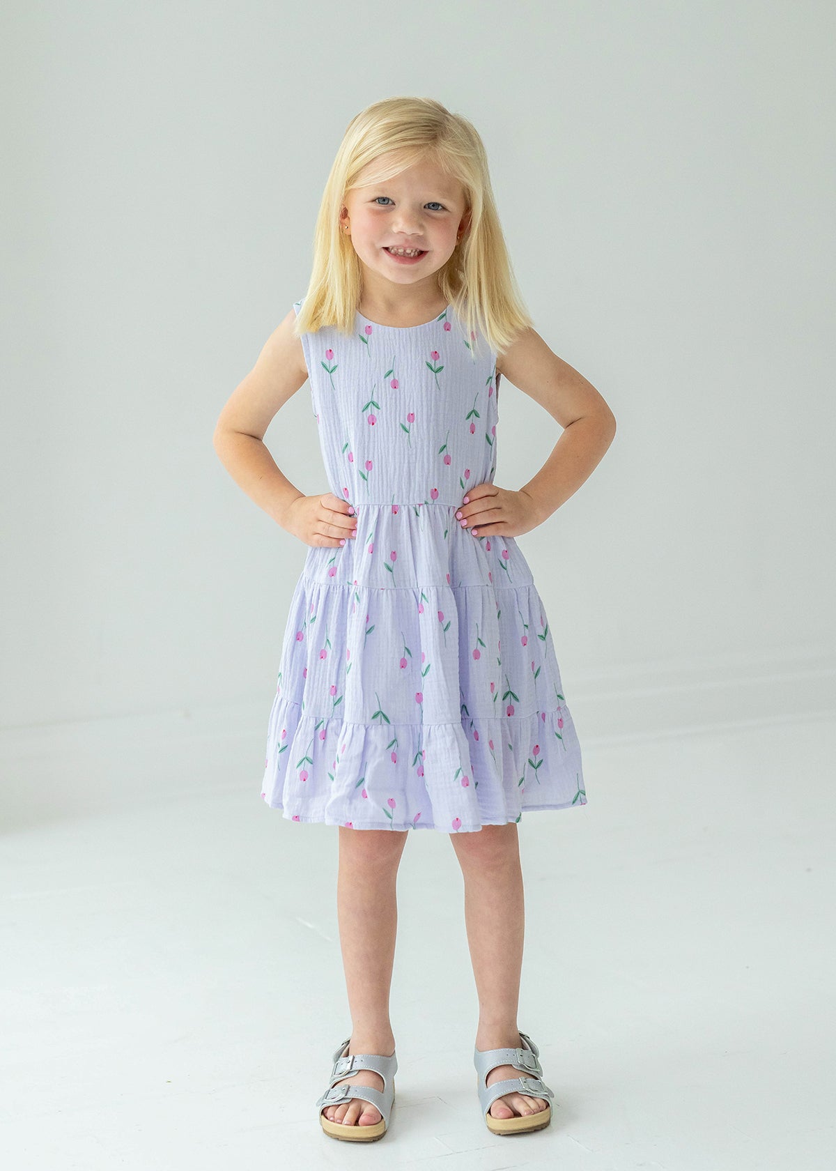 A young blonde girl smiles with her hands on her hips, wearing the Mabel and Honey Tiny Tulip Drop Waist Dress and silver sandals, standing against a plain white background.