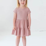 A young blonde girl in pigtails smiles as she models the Josie Dress by Mabel and Honey—a blush pink terry cloth girl's dress—paired with white sandals, against a plain white background.
