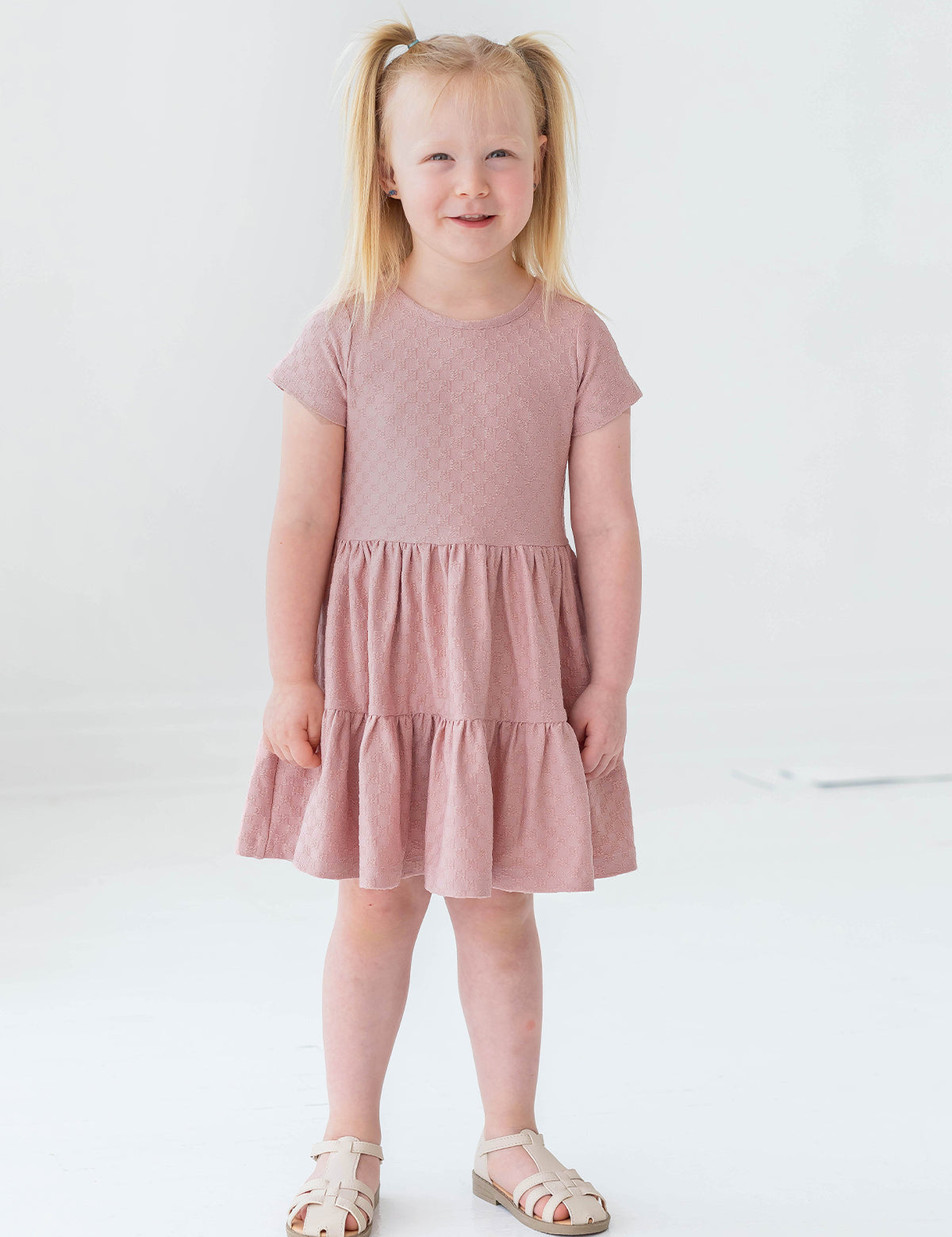 A young blonde girl in pigtails smiles as she models the Josie Dress by Mabel and Honey—a blush pink terry cloth girl's dress—paired with white sandals, against a plain white background.