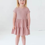 A young blonde girl in pigtails smiles as she models the Josie Dress by Mabel and Honey—a blush pink terry cloth girl's dress—paired with white sandals, against a plain white background.