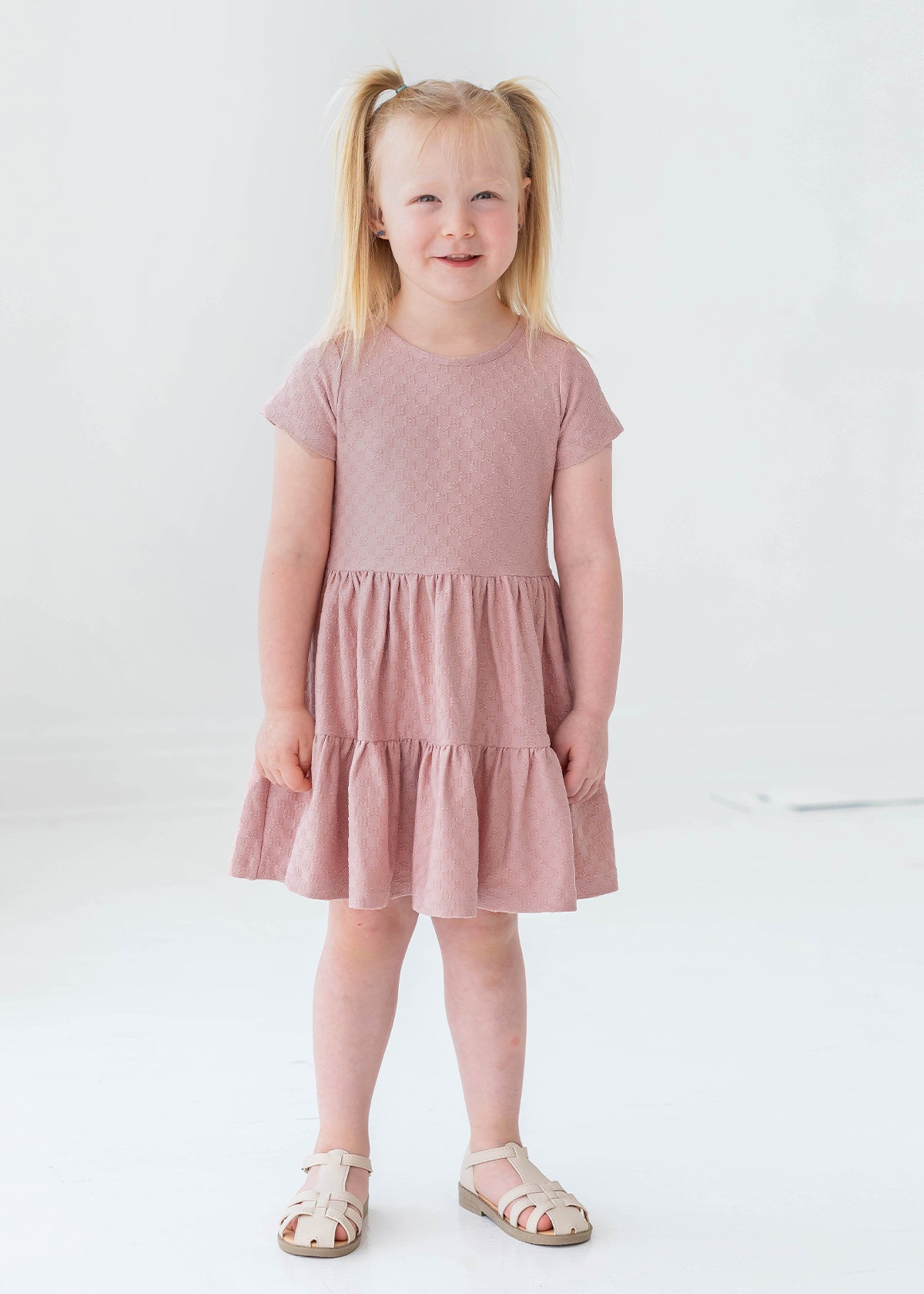 A young blonde girl in pigtails smiles as she models the Josie Dress by Mabel and Honey—a blush pink terry cloth girl's dress—paired with white sandals, against a plain white background.