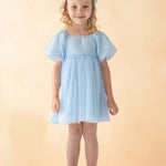 A young girl with blonde curly hair smiles against a light beige background, wearing the Isobella and Chloe Ella Dress in pastel blue with butterfly sleeves, a matching blue hair bow, and white shoes.