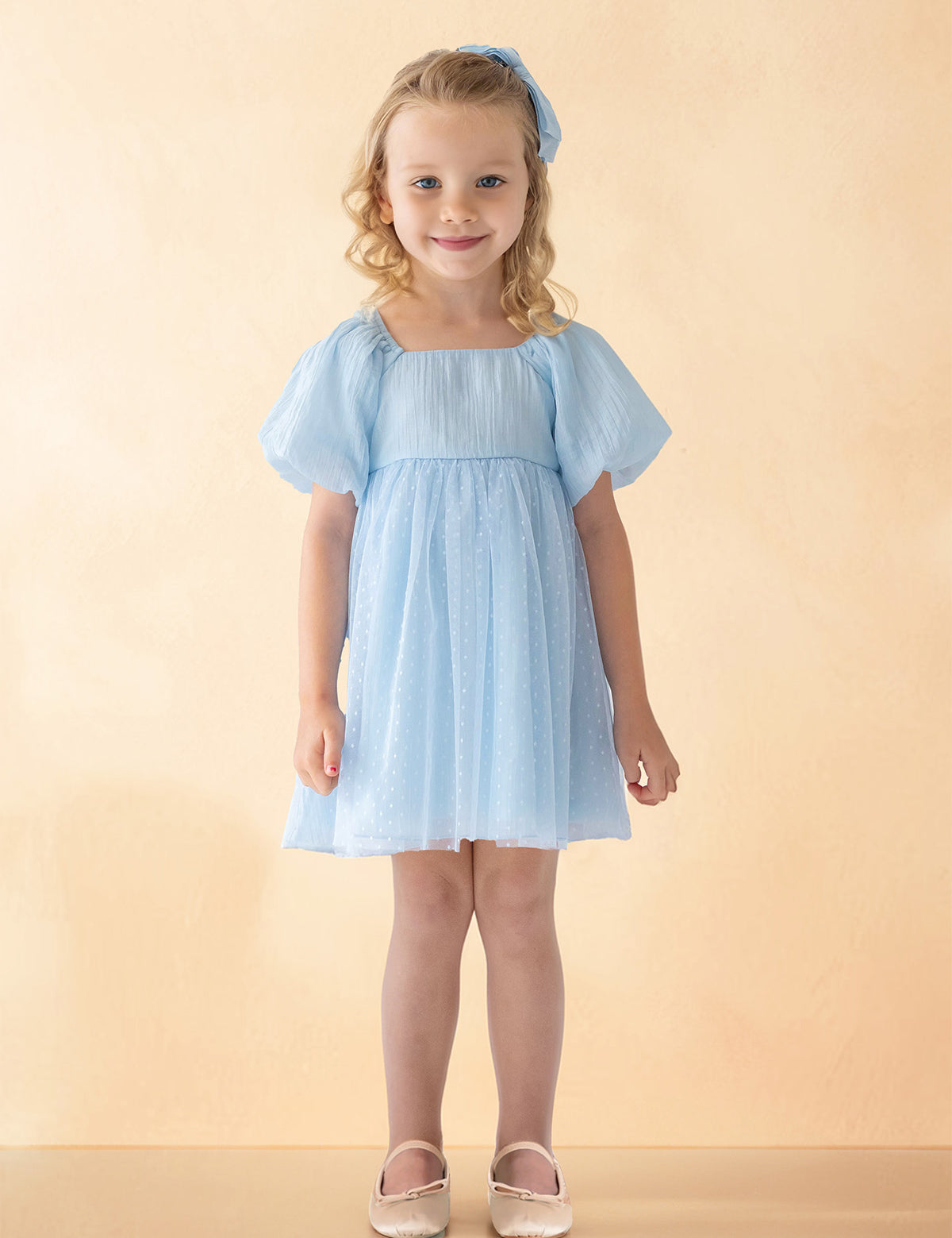 A young girl with blonde curly hair smiles against a light beige background, wearing the Isobella and Chloe Ella Dress in pastel blue with butterfly sleeves, a matching blue hair bow, and white shoes.