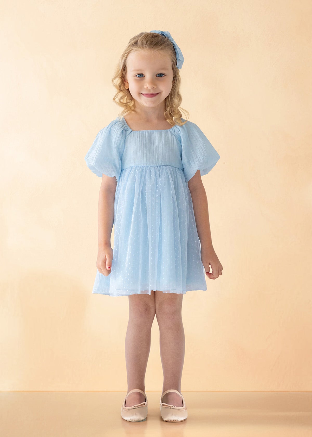 A young girl with blonde curly hair smiles against a light beige background, wearing the Isobella and Chloe Ella Dress in pastel blue with butterfly sleeves, a matching blue hair bow, and white shoes.