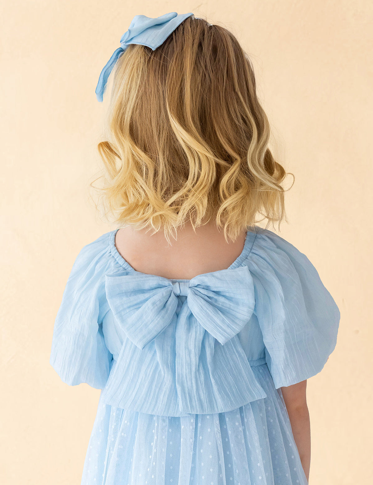 A young girl with blonde, wavy hair is seen from behind wearing the Isobella and Chloe Ella Dress—a pastel blue dress with butterfly sleeves, a large back bow, and a matching hair bow—set against a soft beige background.