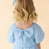A young girl with blonde, wavy hair is seen from behind wearing the Isobella and Chloe Ella Dress—a pastel blue dress with butterfly sleeves, a large back bow, and a matching hair bow—set against a soft beige background.