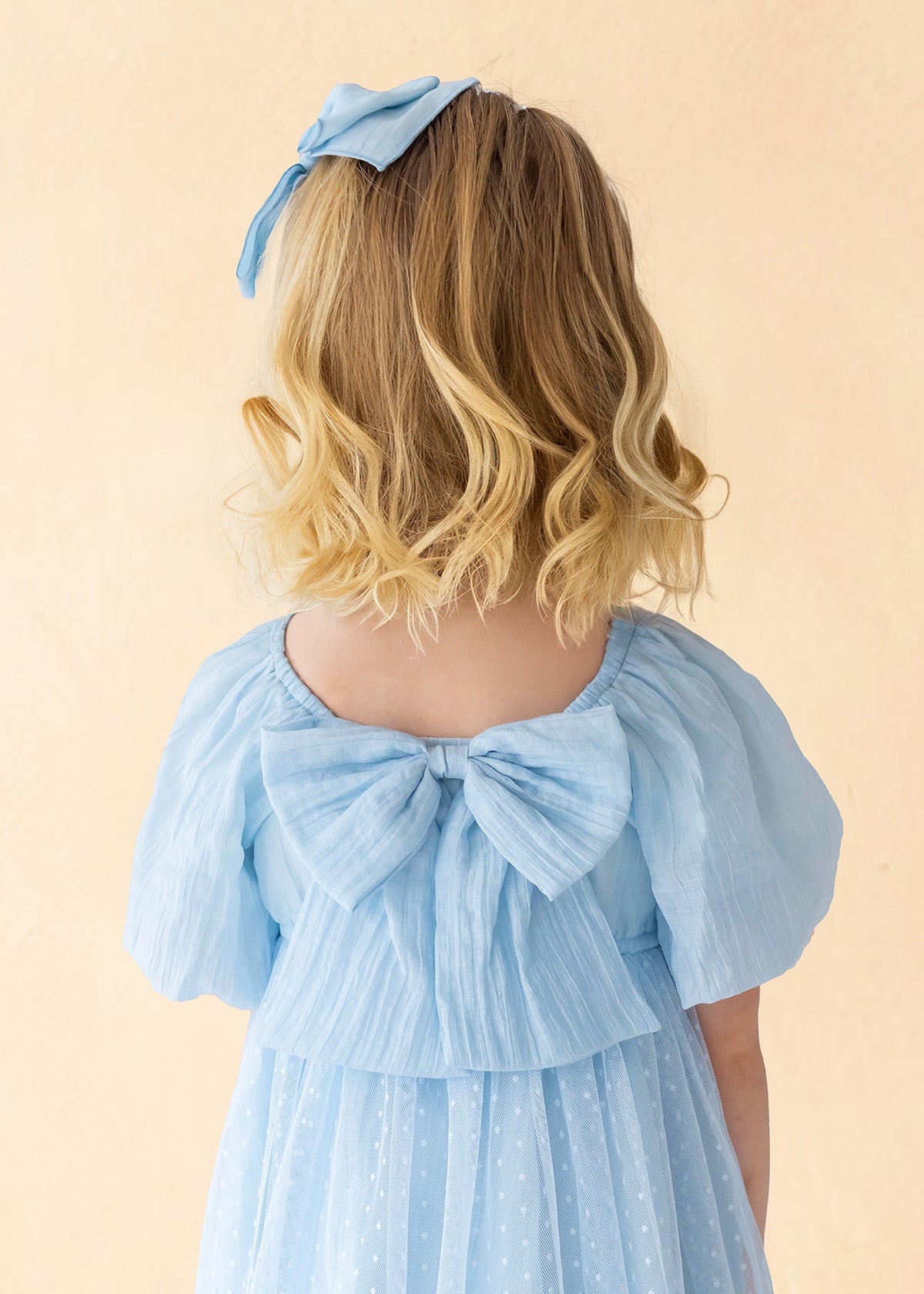 A young girl with blonde, wavy hair is seen from behind wearing the Isobella and Chloe Ella Dress—a pastel blue dress with butterfly sleeves, a large back bow, and a matching hair bow—set against a soft beige background.