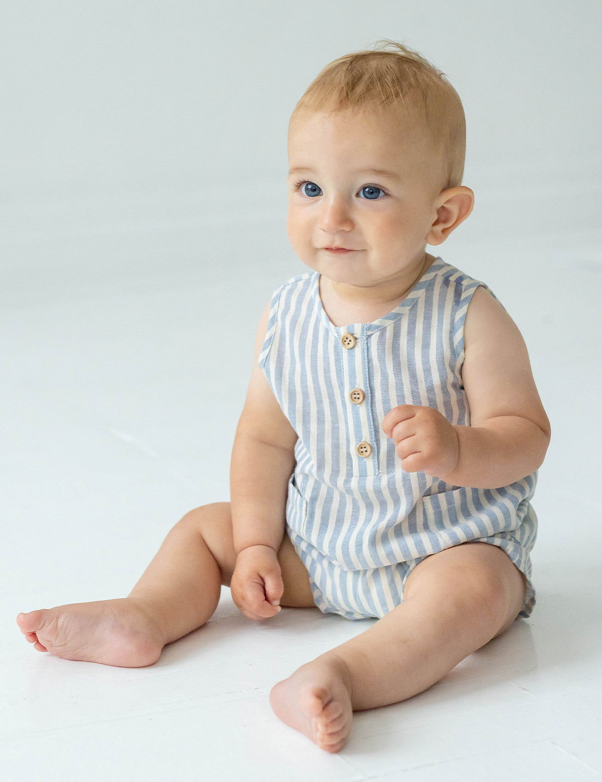 A baby with light hair and blue eyes sits on a white floor, wearing the Beckett and Bear Blake Baby Romper in light blue stripes with wooden buttons. The baby looks slightly upward and smiles softly.