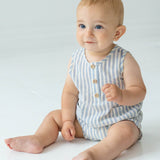 A baby with light hair and blue eyes sits on a white floor, wearing the Beckett and Bear Blake Baby Romper in light blue stripes with wooden buttons. The baby looks slightly upward and smiles softly.