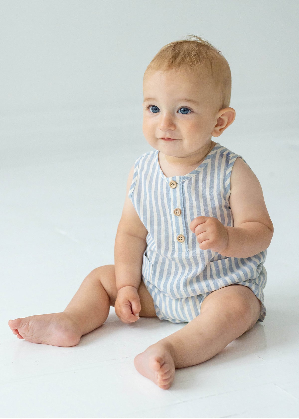 A baby with light hair and blue eyes sits on a white floor, wearing the Beckett and Bear Blake Baby Romper in light blue stripes with wooden buttons. The baby looks slightly upward and smiles softly.