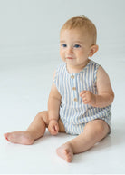 A baby with light hair and blue eyes sits on a white floor, wearing the Beckett and Bear Blake Baby Romper in light blue stripes with wooden buttons. The baby looks slightly upward and smiles softly.
