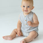A smiling baby with short light hair sits on the floor in the Beckett and Bear Blake Baby Romper, featuring light blue stripes and wooden buttons, looking slightly to the side.