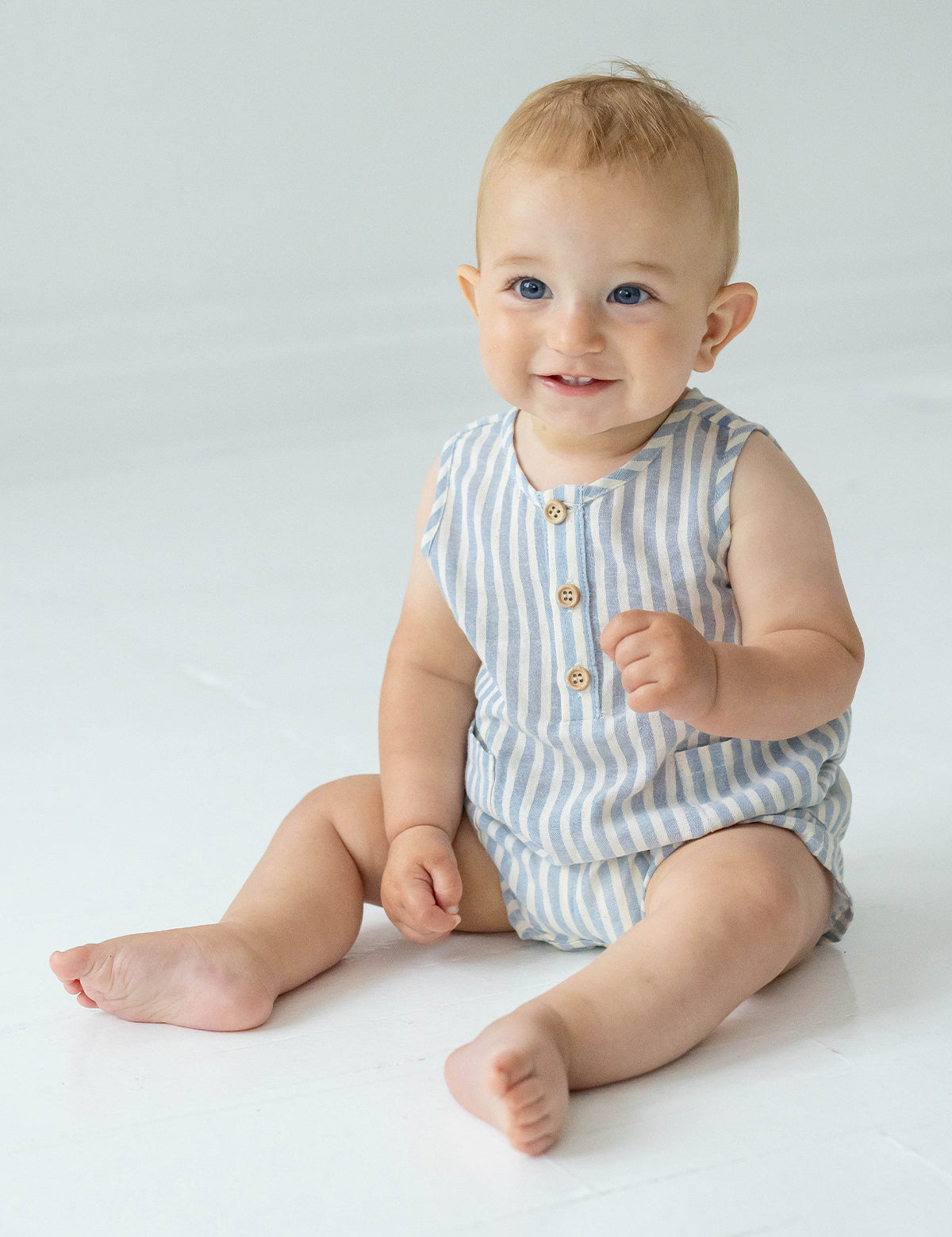 A smiling baby with short light hair sits on the floor in the Beckett and Bear Blake Baby Romper, featuring light blue stripes and wooden buttons, looking slightly to the side.