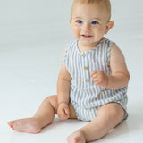 A smiling baby with short light hair sits on the floor in the Beckett and Bear Blake Baby Romper, featuring light blue stripes and wooden buttons, looking slightly to the side.