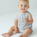 A smiling baby with blue eyes and light hair sits barefoot on the floor in a Beckett and Bear Blake Baby Romper, looking at the camera against a plain, light background.