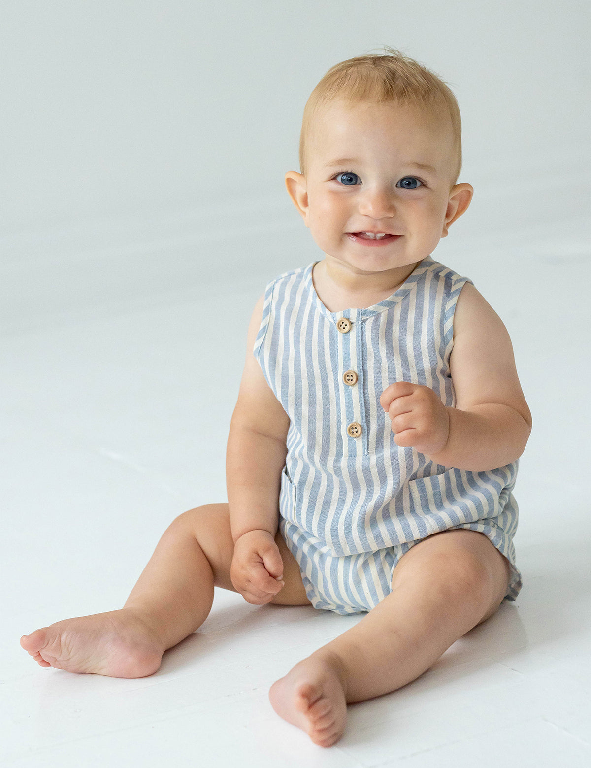 A smiling baby with blue eyes and light hair sits barefoot on the floor in a Beckett and Bear Blake Baby Romper, looking at the camera against a plain, light background.