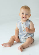 A smiling baby with blue eyes and light hair sits barefoot on the floor in a Beckett and Bear Blake Baby Romper, looking at the camera against a plain, light background.