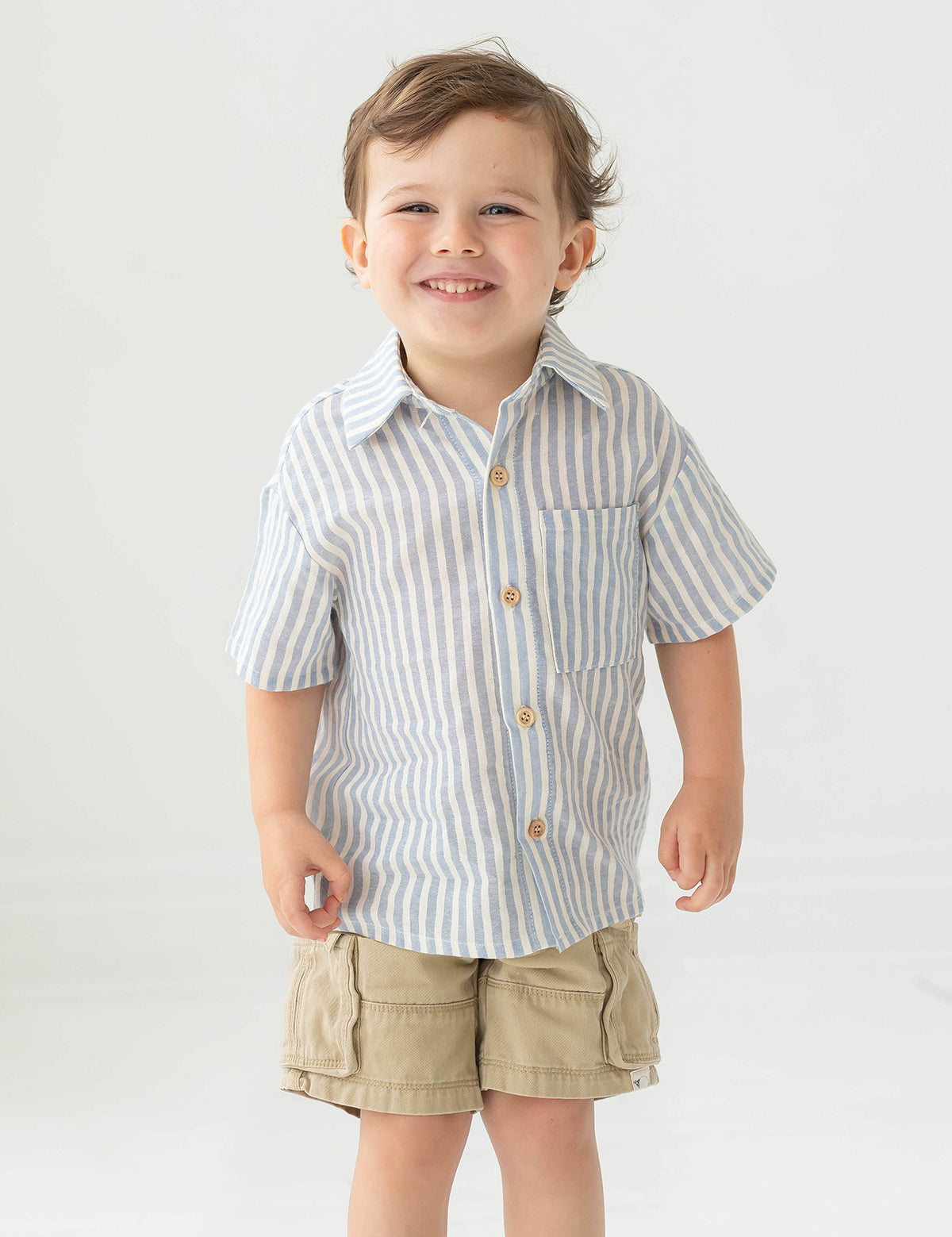 A young boy with short brown hair smiles while wearing the Blake Top from Beckett and Bear, a light blue striped, 100% cotton shirt, paired with beige cargo shorts, standing against a plain light background.