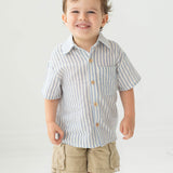 A young boy with short brown hair smiles while wearing the Blake Top from Beckett and Bear, a light blue striped, 100% cotton shirt, paired with beige cargo shorts, standing against a plain light background.