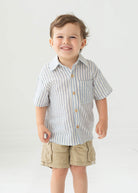 A young boy with short brown hair smiles while wearing the Blake Top from Beckett and Bear, a light blue striped, 100% cotton shirt, paired with beige cargo shorts, standing against a plain light background.