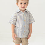 A young boy with short light brown hair stands against a plain background, smiling slightly in the Beckett and Bear Blake Top, a 100% cotton light blue striped short-sleeve button-up shirt, paired with beige shorts.