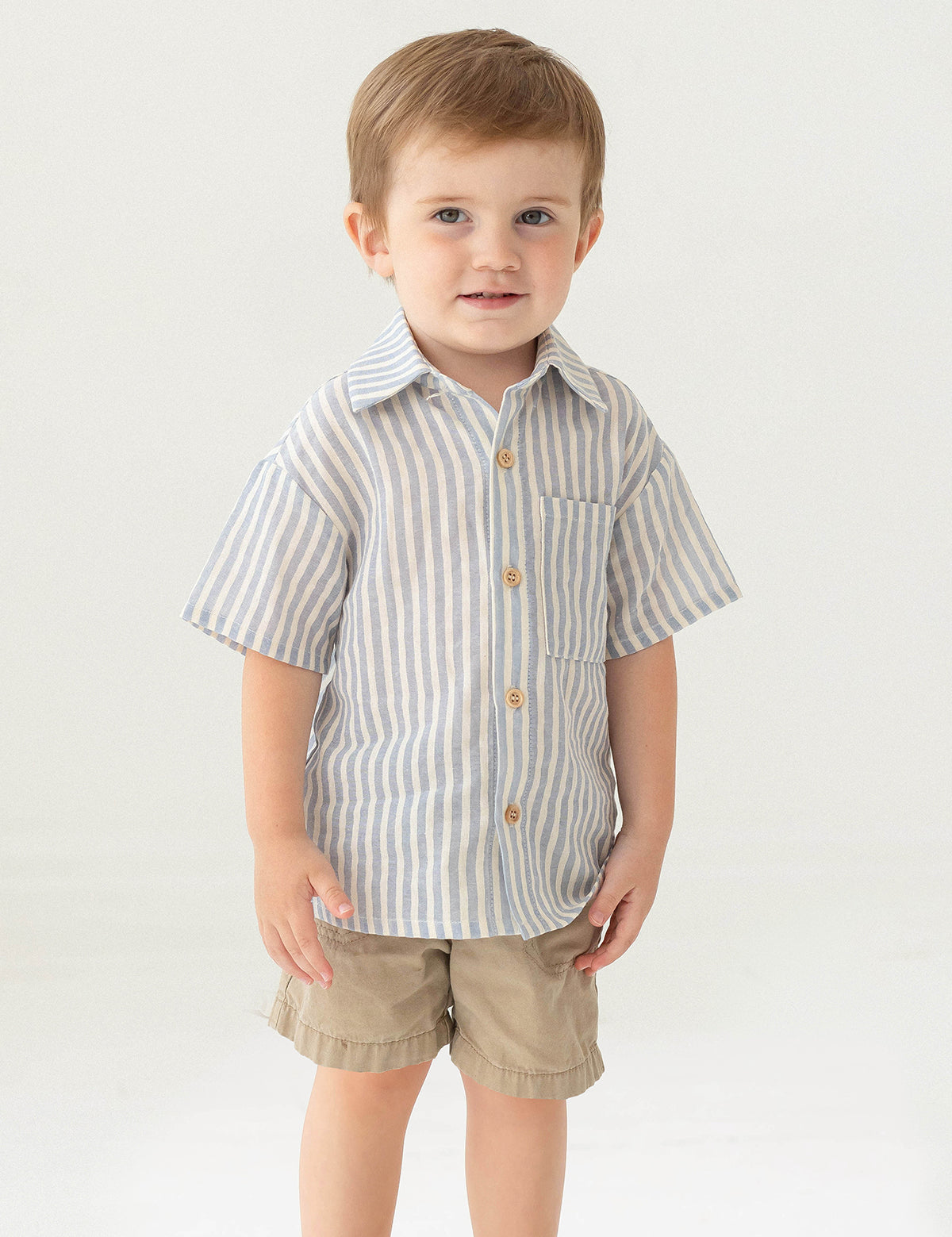 A young boy with short light brown hair stands against a plain background, smiling slightly in the Beckett and Bear Blake Top, a 100% cotton light blue striped short-sleeve button-up shirt, paired with beige shorts.