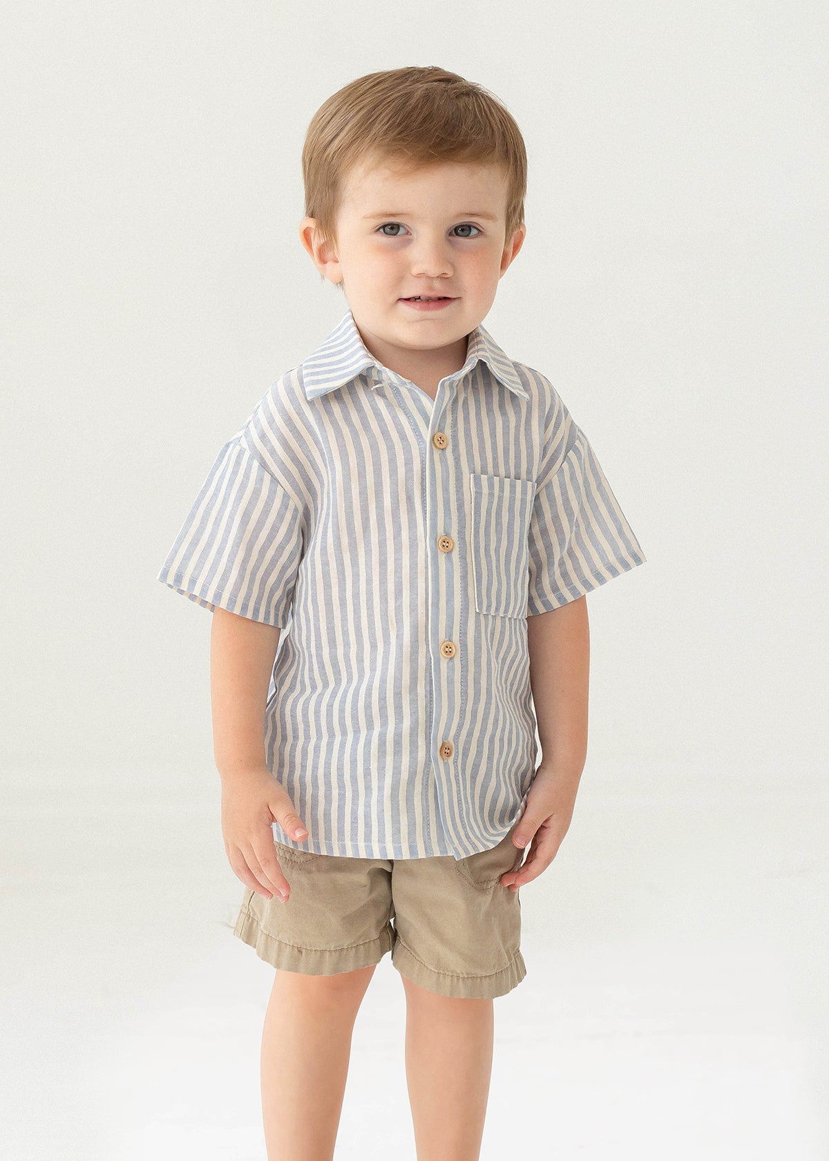 A young boy with short light brown hair stands against a plain background, smiling slightly in the Beckett and Bear Blake Top, a 100% cotton light blue striped short-sleeve button-up shirt, paired with beige shorts.