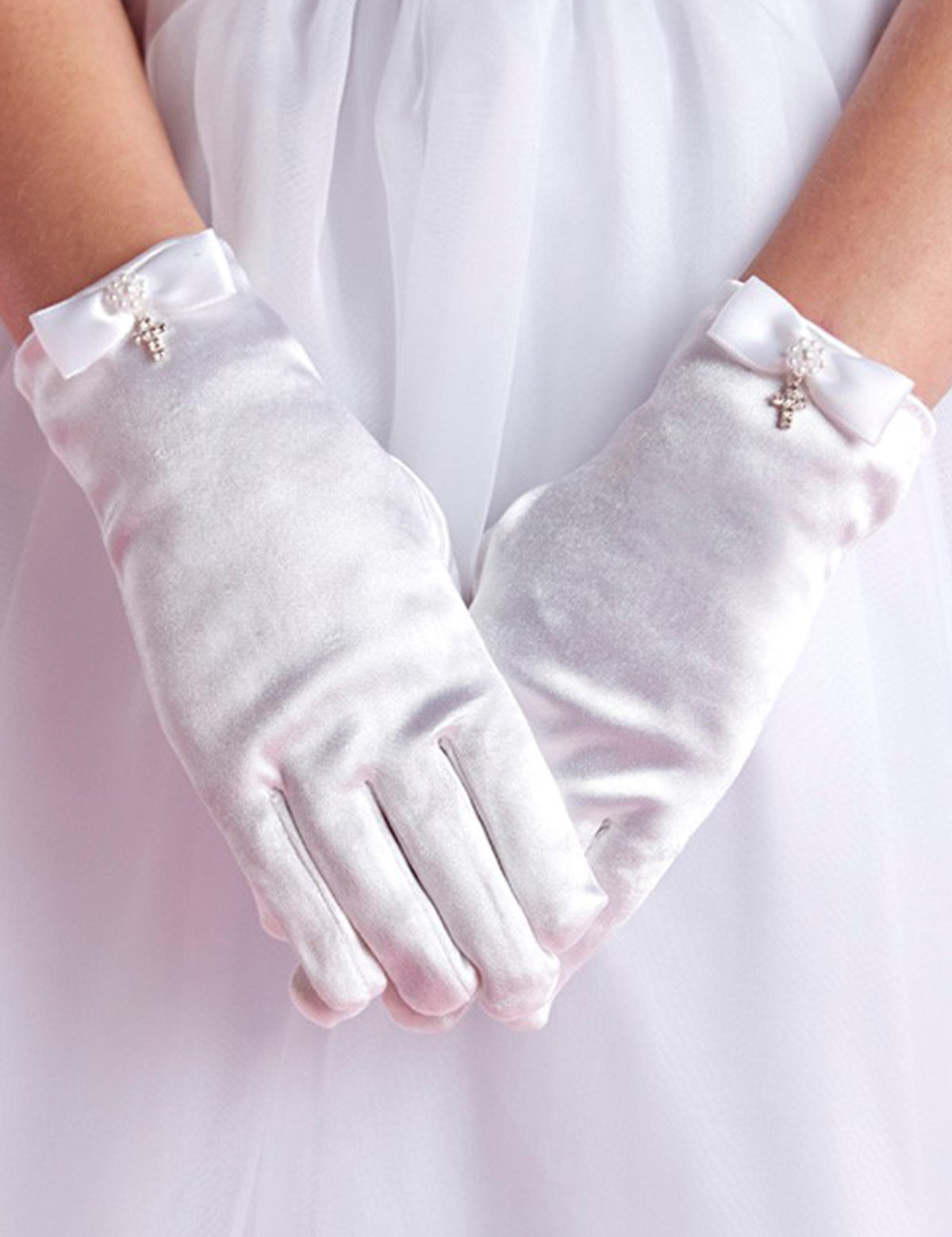 A person wears Lauren Marie Rhinestones & Pearl Cross Gloves, detailed with small bows and a crystal cross charm, standing with hands clasped in front of a white, flowy dress.