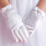 A person wears Lauren Marie Rhinestones & Pearl Cross Gloves, detailed with small bows and a crystal cross charm, standing with hands clasped in front of a white, flowy dress.