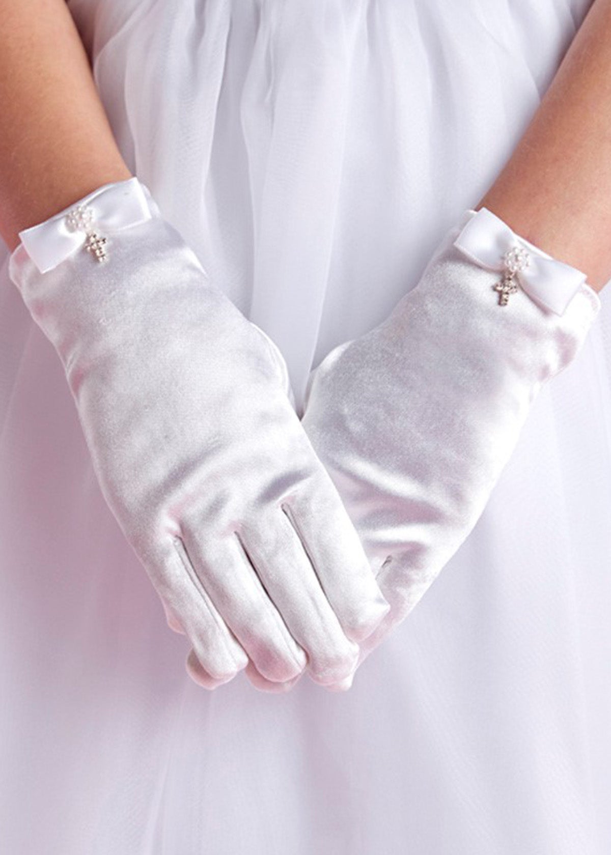 A person wears Lauren Marie Rhinestones & Pearl Cross Gloves, detailed with small bows and a crystal cross charm, standing with hands clasped in front of a white, flowy dress.