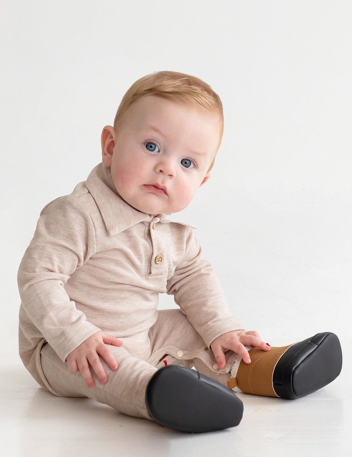 A baby with light hair and blue eyes sits on the floor in a beige Millie Brothers Romper by Beckett and Bear, featuring a collared neckline, long sleeves, and black shoes. The child gazes to the side against a plain white background.