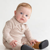 A baby with light hair and blue eyes sits on the floor in a beige Millie Brothers Romper by Beckett and Bear, featuring a collared neckline, long sleeves, and black shoes. The child gazes to the side against a plain white background.
