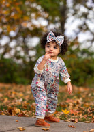 A toddler with curly hair wears the Mabel and Honey Melody Romper and a matching bow headband, standing outdoors on a leaf-strewn path with autumn foliage in the background.