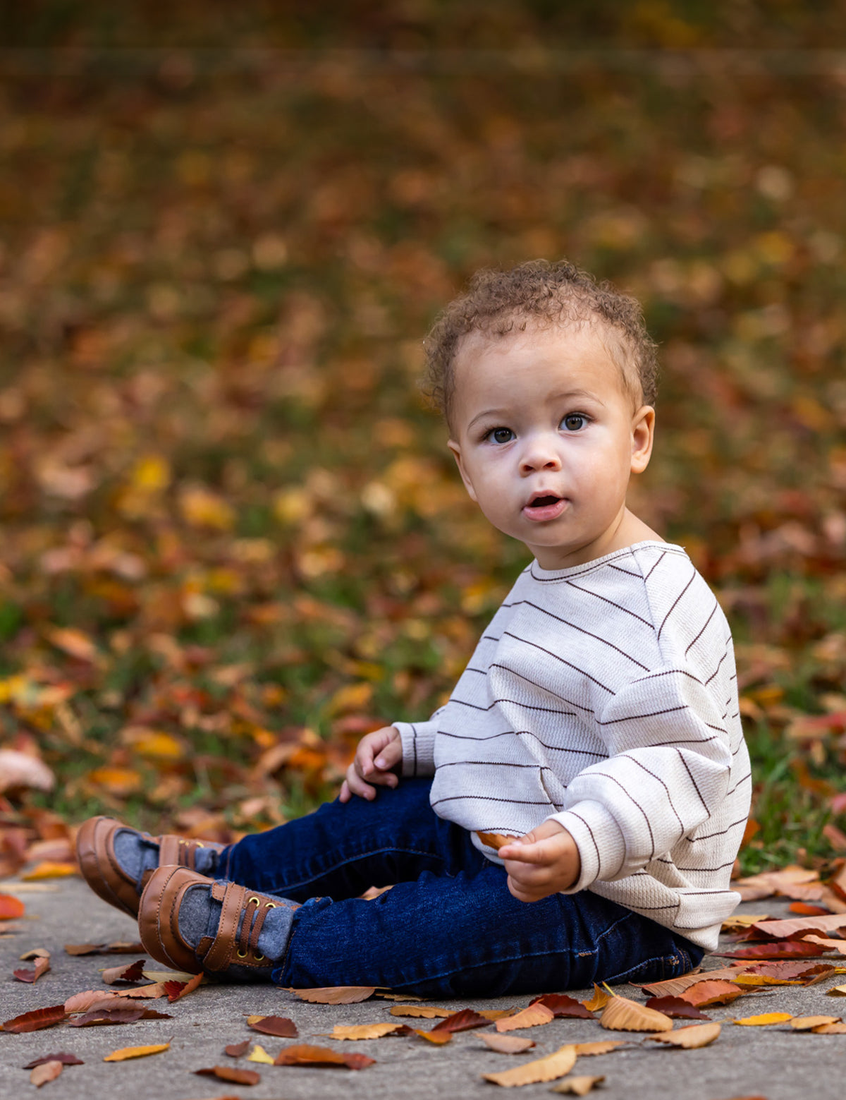 A young child with curly hair sits on a sidewalk among autumn leaves, wearing the Beckett and Bear Brothers Top, blue jeans, and brown shoes, looking curiously toward the camera.