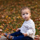 A young child with curly hair sits on a sidewalk among autumn leaves, wearing the Beckett and Bear Brothers Top, blue jeans, and brown shoes, looking curiously toward the camera.