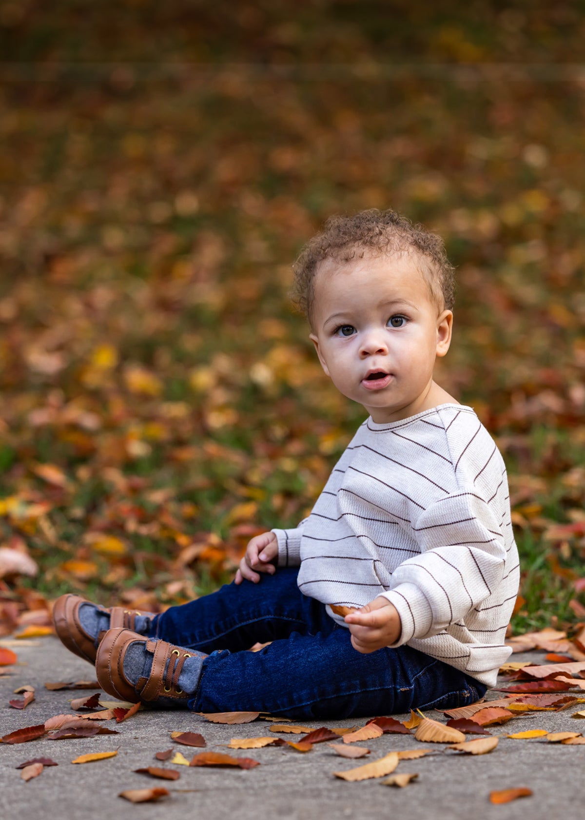 A young child with curly hair sits on a sidewalk among autumn leaves, wearing the Beckett and Bear Brothers Top, blue jeans, and brown shoes, looking curiously toward the camera.