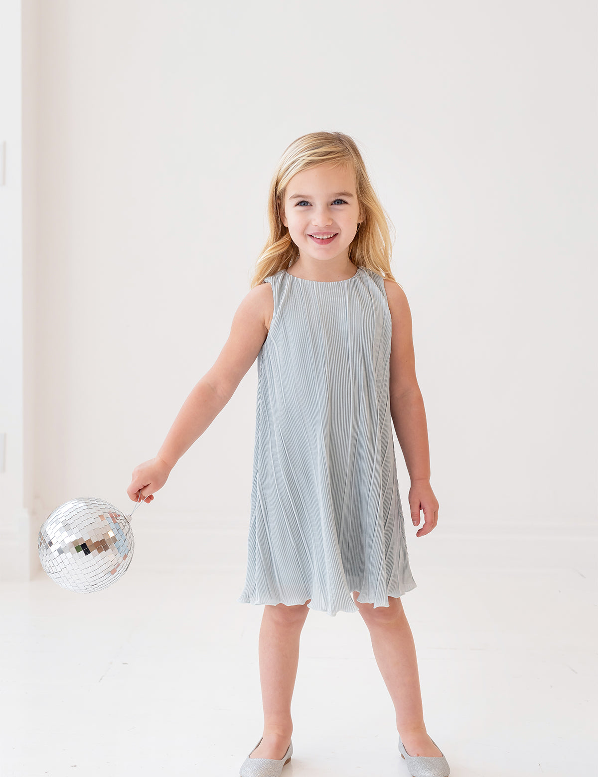 A young girl with blonde hair smiles while holding a small disco ball, dressed in the Isobella and Chloe Wintermelon Dress—a light blue pleated style with matching flats—perfect for any girls’ dress collection against a white background.