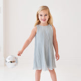 A young girl with blonde hair smiles while holding a small disco ball, dressed in the Isobella and Chloe Wintermelon Dress—a light blue pleated style with matching flats—perfect for any girls’ dress collection against a white background.