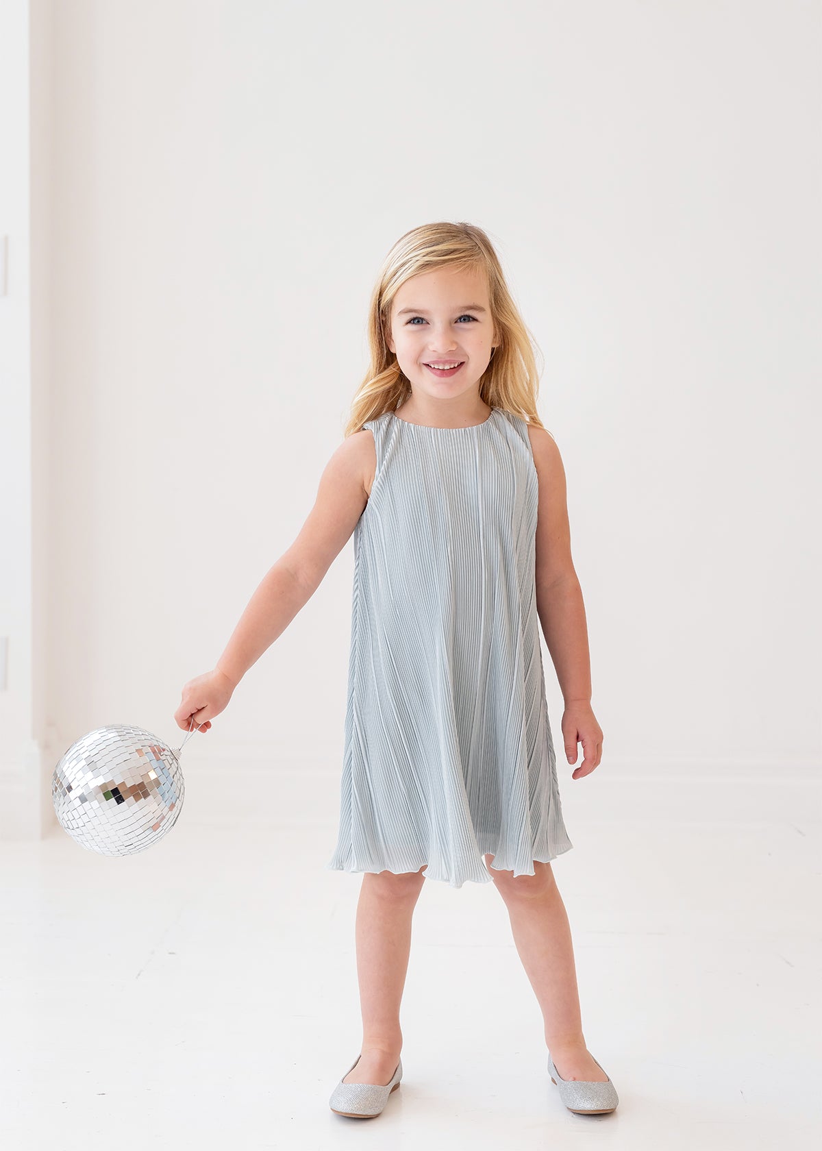 A young girl with blonde hair smiles while holding a small disco ball, dressed in the Isobella and Chloe Wintermelon Dress—a light blue pleated style with matching flats—perfect for any girls’ dress collection against a white background.