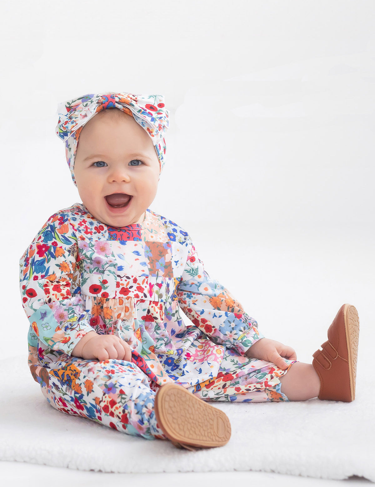 A smiling baby sits on a white blanket, wearing the Flower Arrangement Romper by Mabel and Honey with brown shoes and a matching headband, against a plain white background.