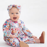 A smiling baby sits on a white blanket, wearing the Flower Arrangement Romper by Mabel and Honey with brown shoes and a matching headband, against a plain white background.