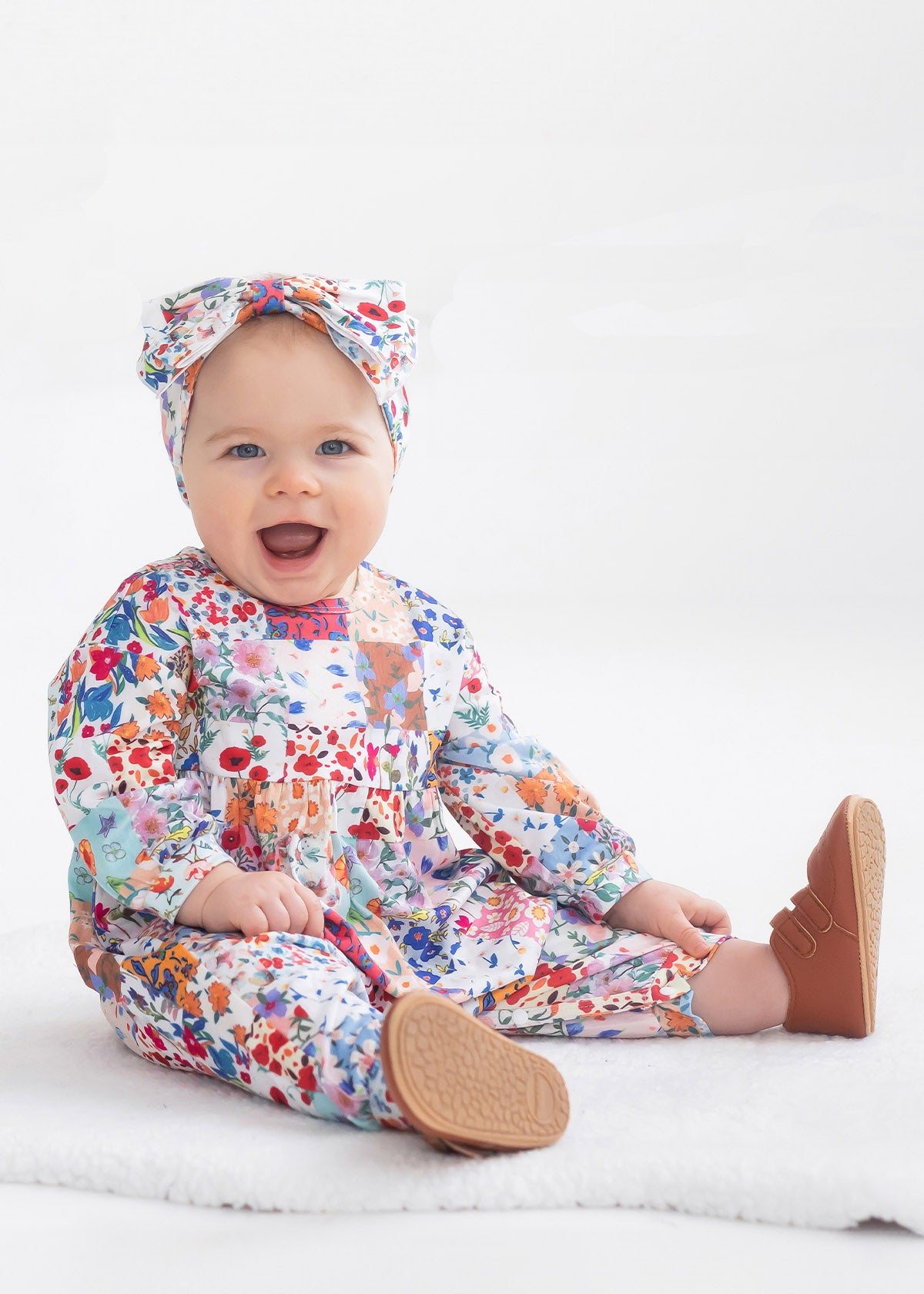 A smiling baby sits on a white blanket, wearing the Flower Arrangement Romper by Mabel and Honey with brown shoes and a matching headband, against a plain white background.