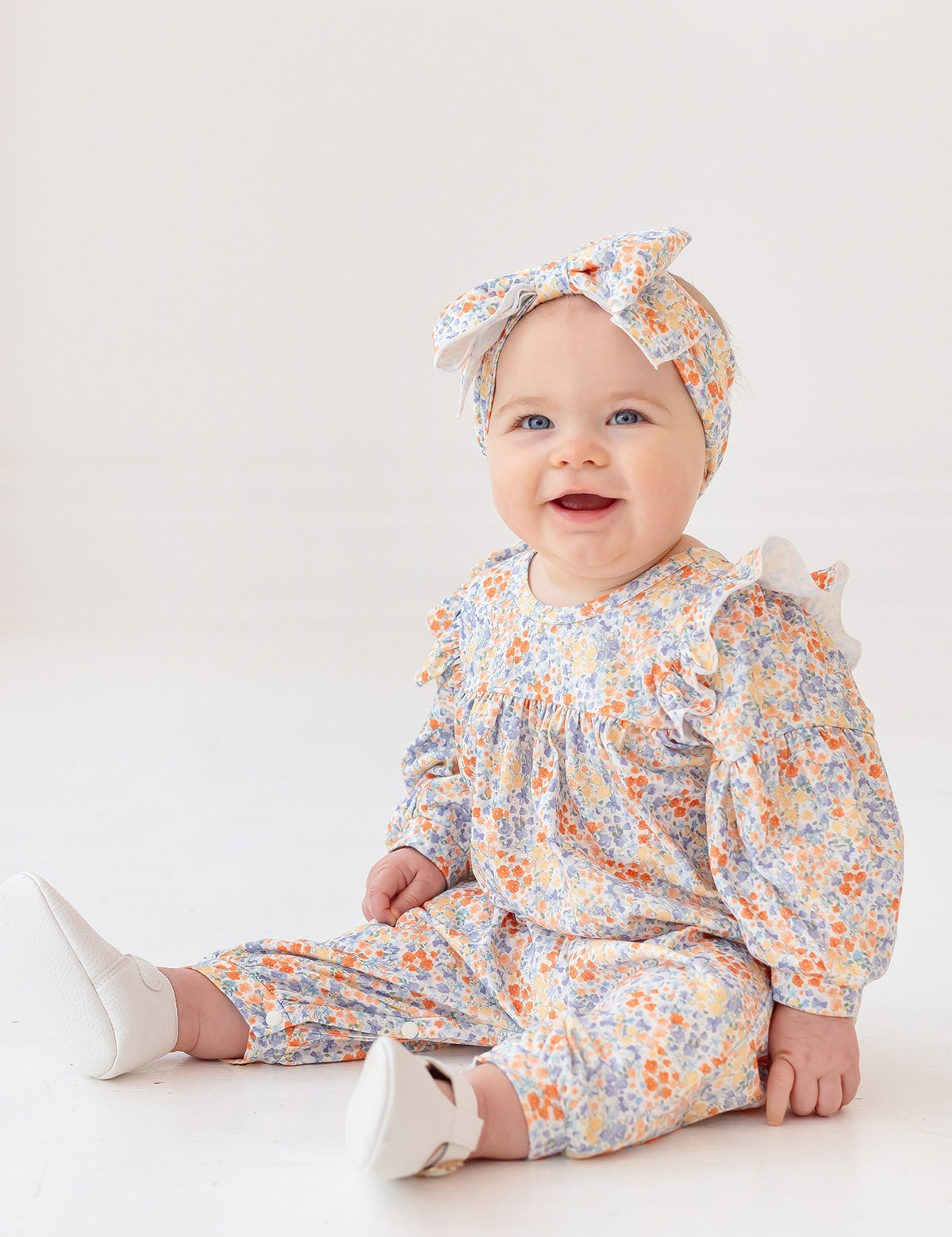 A smiling baby sits on the floor in the Mabel and Honey Darling Orchard Romper with a matching headband and white shoes, against a plain light background.