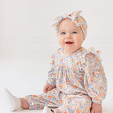 A smiling baby sits on the floor in the Mabel and Honey Darling Orchard Romper with a matching headband and white shoes, against a plain light background.
