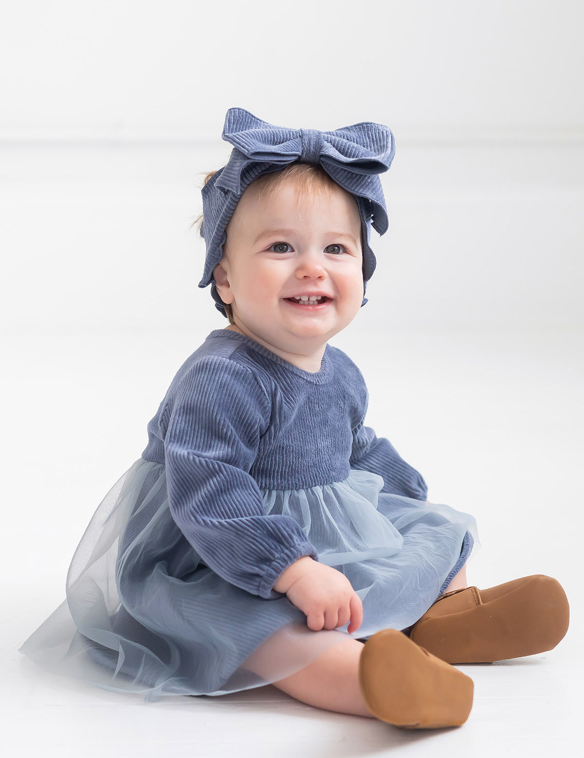 A smiling baby with light hair sits on the floor in a Mabel and Honey Skylar Blue Suede Dress, styled with a tulle skirt, corduroy bodice, a matching blue bow headband, and brown shoes against a plain white background.